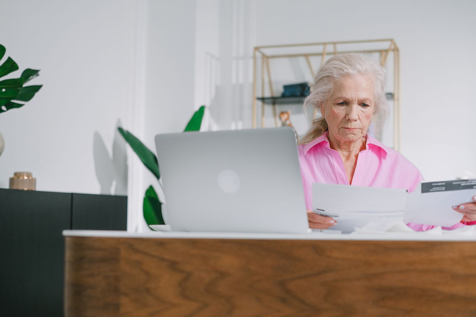 An older woman reviewing papers beside her laptop at home.