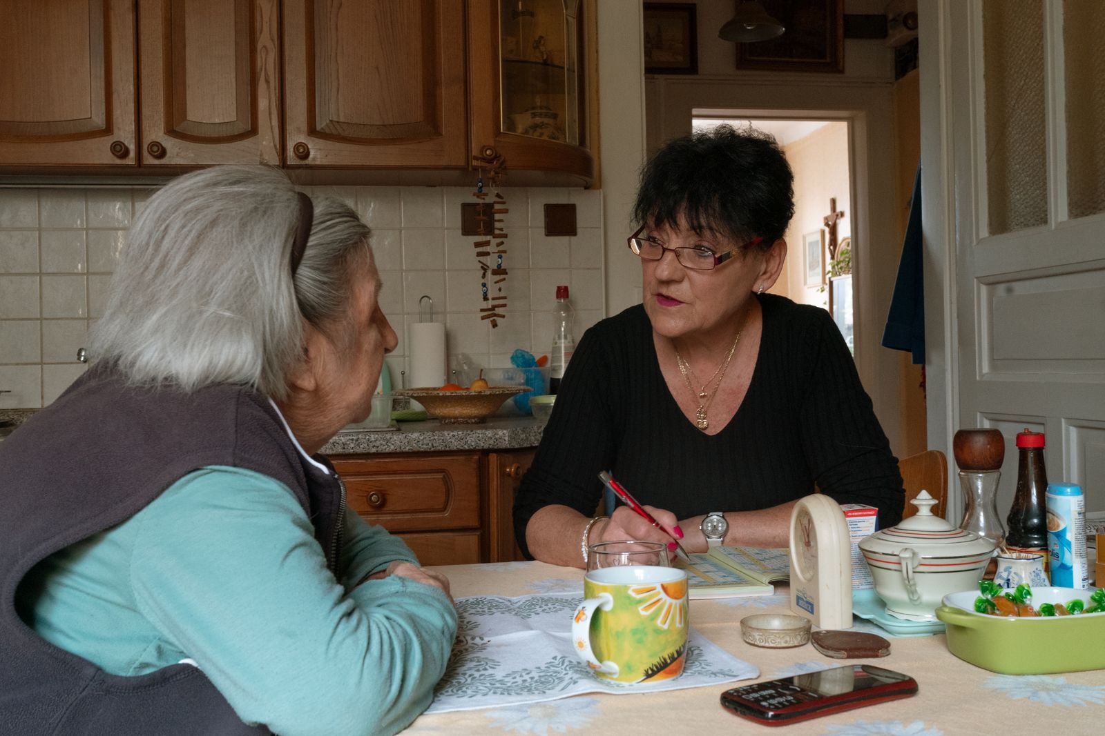 An older woman and a caregiver talking across a kitchen table.